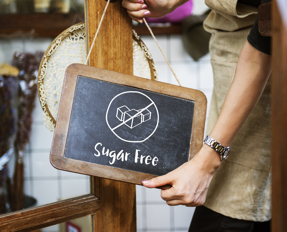 A person in an apron holds a wooden sign that reads "Sugar Free," featuring a graphic of sugar cubes with a line through them, in a cozy shop setting.