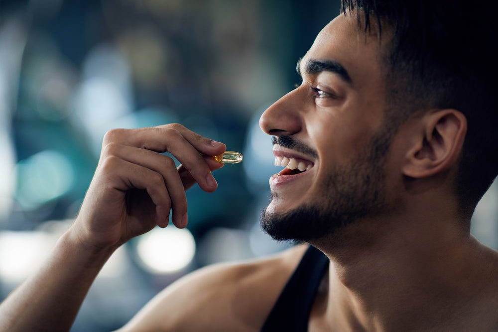 A smiling man holds a nutritional supplement capsule in his hand, preparing to take it, with a blurred gym setting in the background, emphasizing the importance of supplements after weight loss surgery.