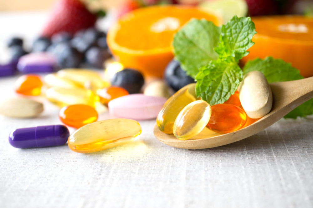 A wooden spoon holds various colorful dietary supplements, including gel capsules and tablets, surrounded by fresh fruits like strawberries, blueberries, and orange slices on a light background.
