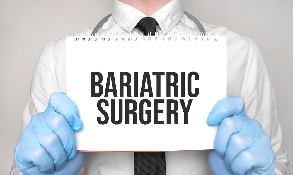 A person in a white shirt and tie, wearing blue gloves, holds a sign that prominently displays the words "BARIATRIC SURGERY" against a neutral background.