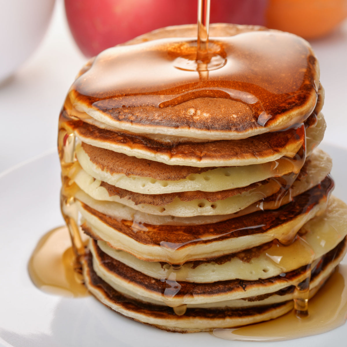 A stack of fluffy pancakes drizzled with syrup sits on a white plate, with apples and an orange blurred in the background, suggesting a cozy breakfast setting.