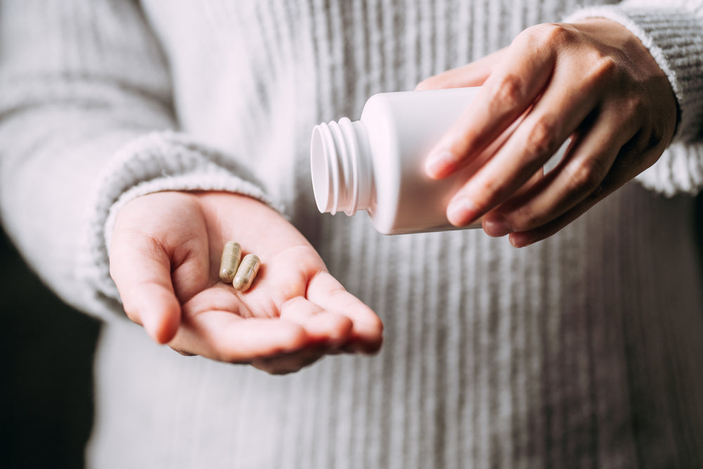 A person in a cozy sweater is pouring vitamins from a white bottle into their palm, highlighting the importance of vitamin intake for bariatric patients.