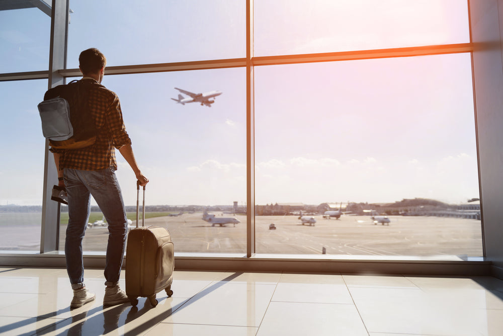 A traveler stands in an airport terminal, looking out a large window at an airplane taking off, with a suitcase in one hand and a backpack on their back.