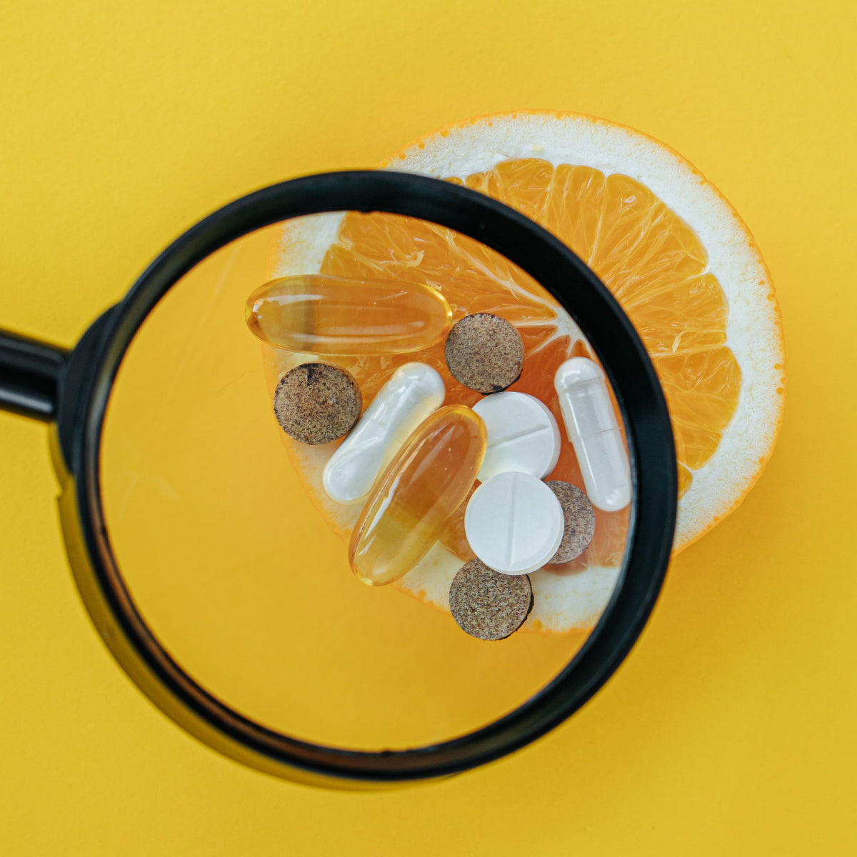 A magnifying glass focuses on various vitamin supplements placed on a slice of orange, set against a bright yellow background, highlighting their importance for nutrition after bariatric surgery.