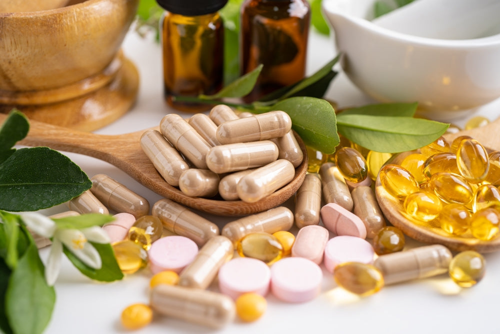 A wooden spoon holds beige capsules among various vitamins and supplements, with green leaves and small bottles in the background, suggesting a focus on health and nutrition.