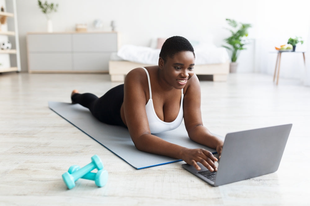 A woman with short hair smiles while lying on a yoga mat, using a laptop in a bright, modern room with plants and a dumbbell nearby, focusing on her weight loss journey.