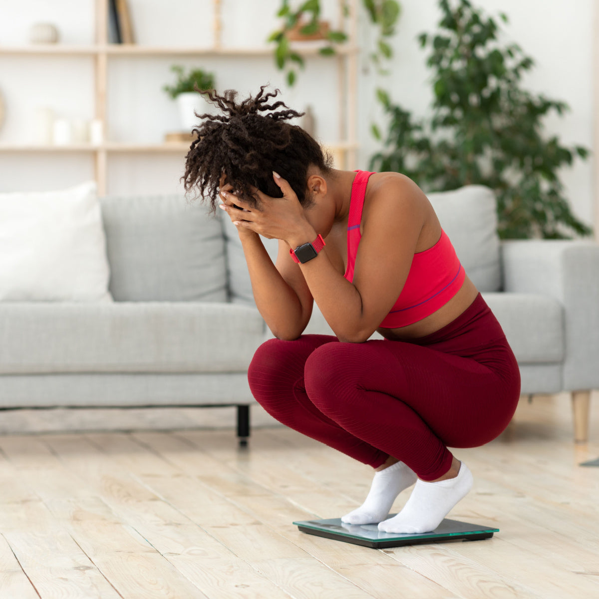 A woman in a pink sports bra and red leggings squats on a scale with her head in her hands, expressing frustration in a bright, modern living room with a couch and plants.