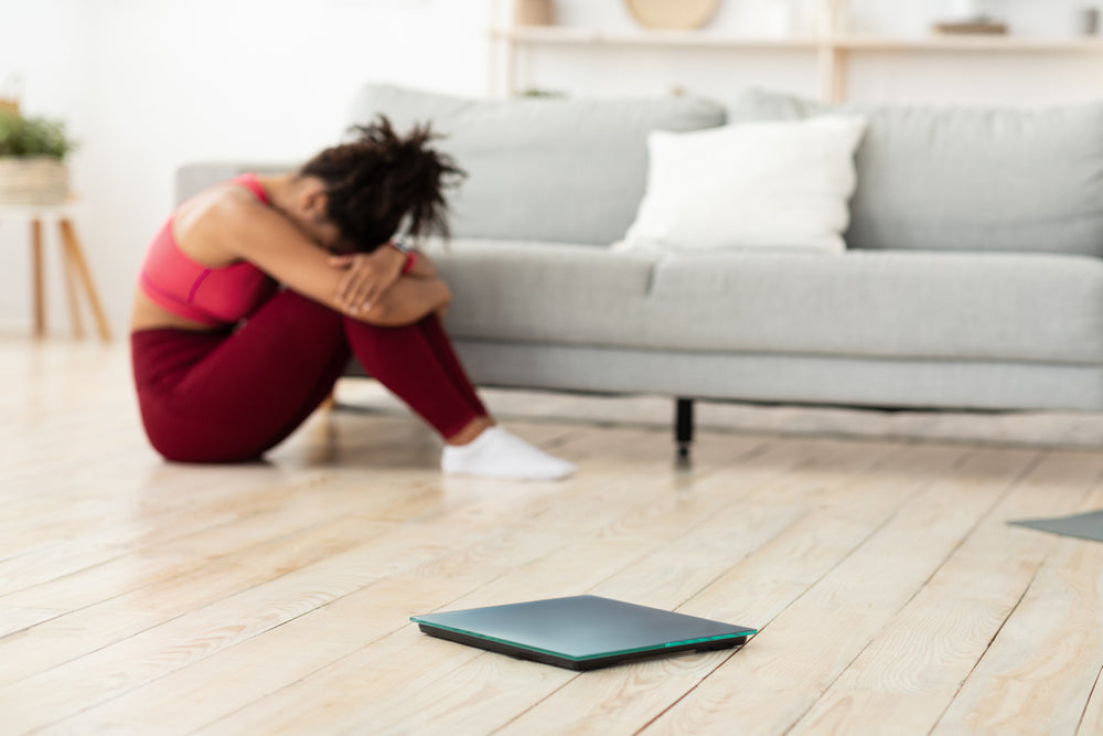 A woman in a pink sports bra and red leggings sits on the floor with her knees hugged to her chest, looking distressed, while a scale lies nearby in a bright, modern living room.