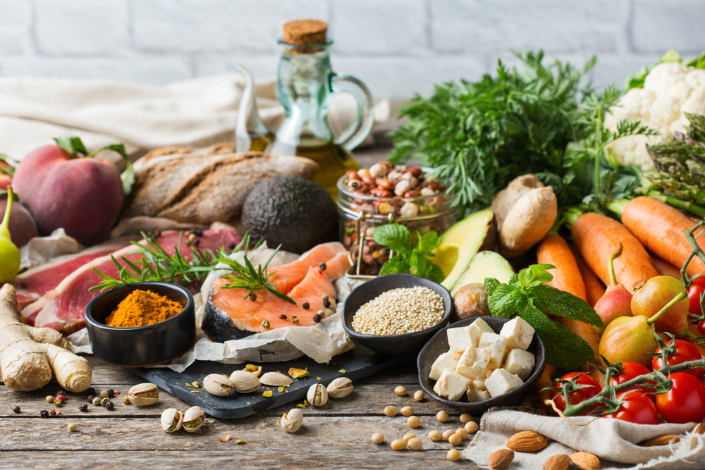 A variety of fresh, healthy foods including salmon, vegetables, fruits, nuts, and spices are artfully arranged on a wooden table, promoting a balanced diet and healthy eating.