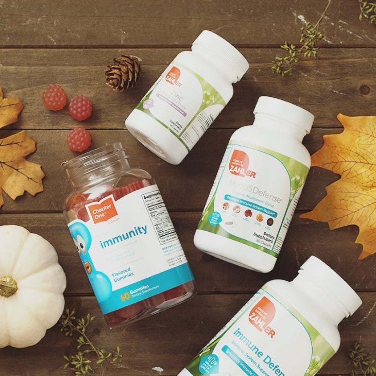 Four bottles of Zahler dietary supplements, including immunity gummies and mushroom blends, are arranged on a wooden surface with autumn leaves and berries, highlighting health and wellness products.