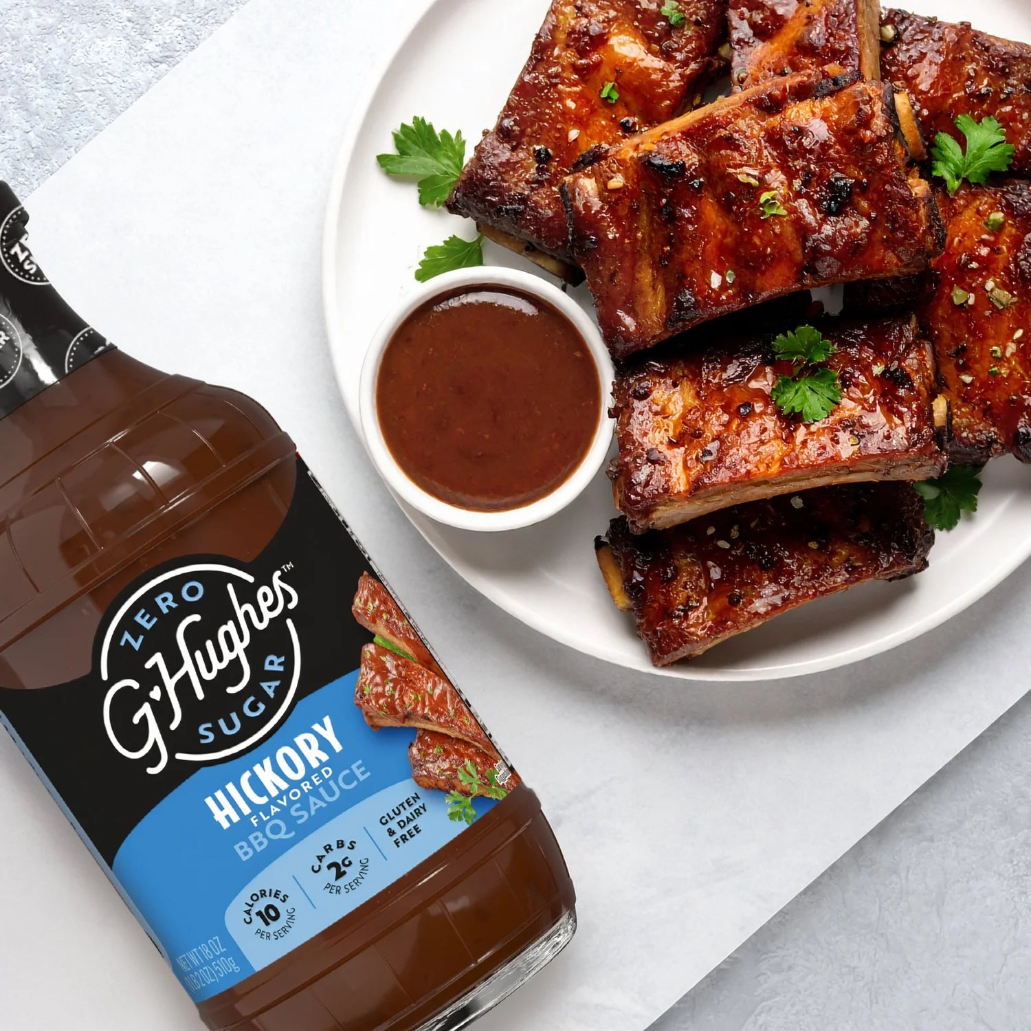 A bottle of G. Hughes Zero Sugar Hickory BBQ Sauce is displayed next to a plate of glazed ribs garnished with parsley and served with a small bowl of BBQ sauce.
