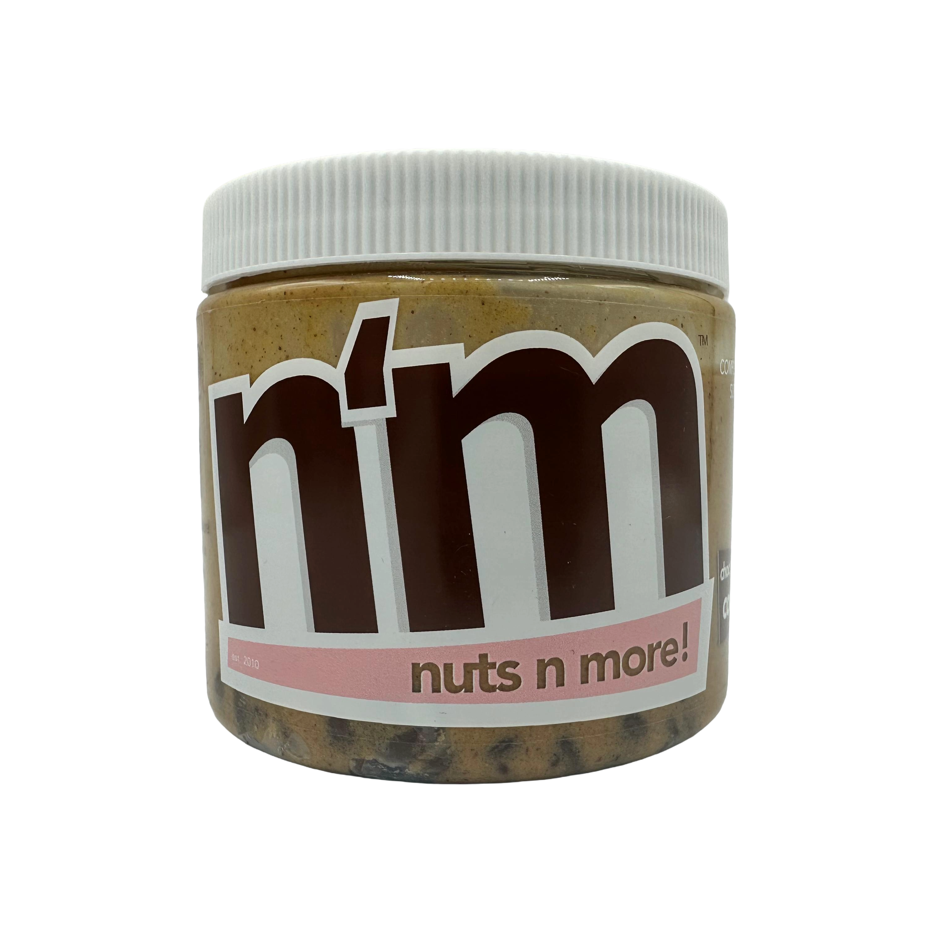 A clear jar of nut butter labeled "n'm" with a white lid, featuring a bold brown logo and the tagline "nuts n more!" on a pink band, set against a black background.
