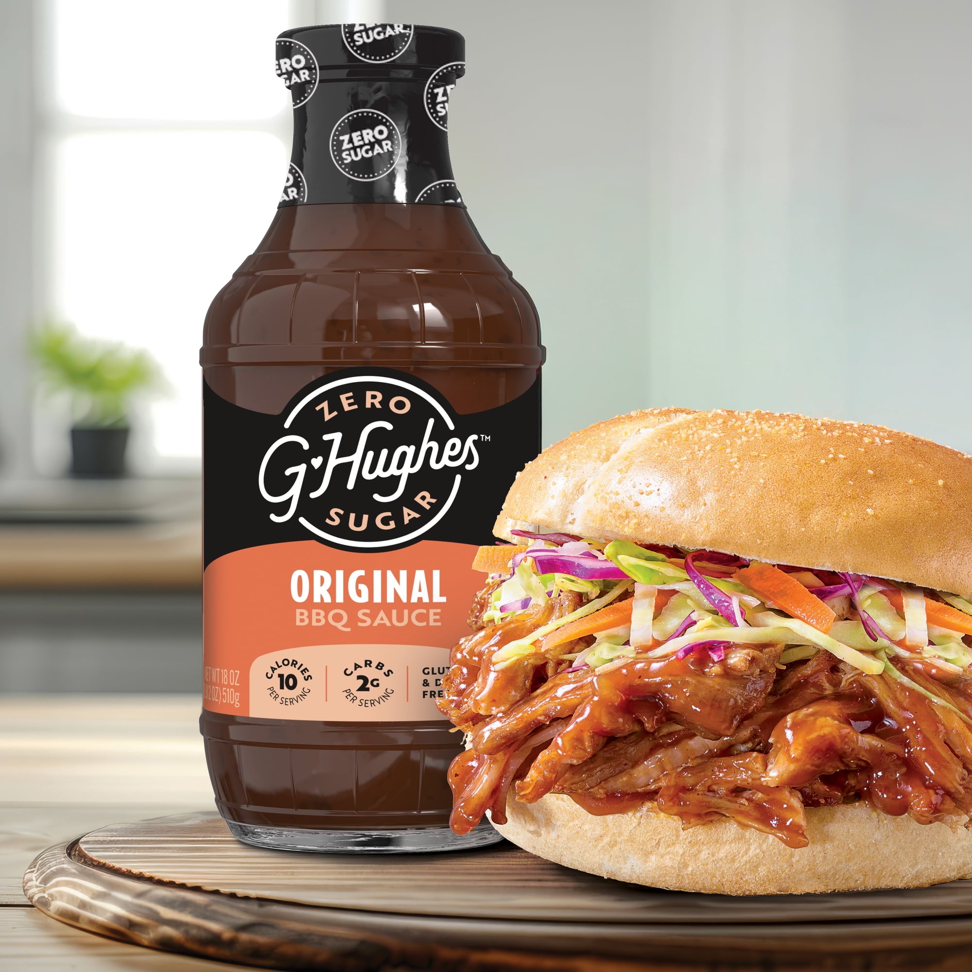A bottle of G. Hughes Zero Sugar Original BBQ Sauce stands next to a piled-high pulled pork sandwich topped with colorful slaw, set on a wooden surface in a bright kitchen.