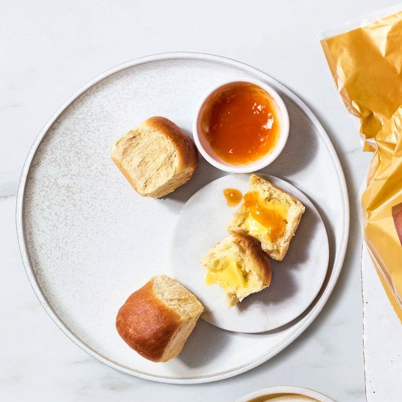 A round plate holds three soft, golden-brown rolls, one cut open with butter and orange jam, alongside a small bowl of jam, set against a light marble background.