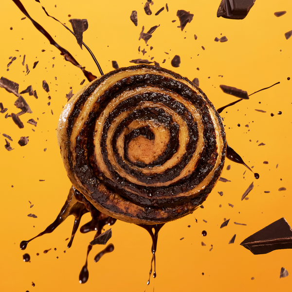 A chocolate swirl pastry is suspended mid-air, surrounded by splashes of melted chocolate and flying chocolate pieces, against a vibrant orange background.