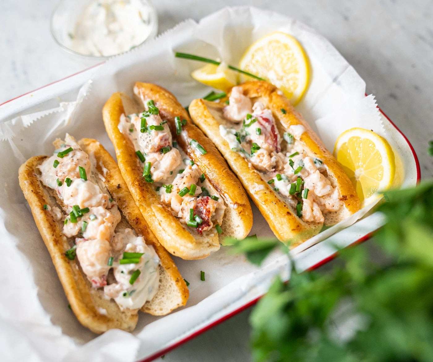 Three toasted rolls filled with creamy lobster salad topped with chives, served with lemon slices, and accompanied by a small bowl of sauce, all presented on a white tray.