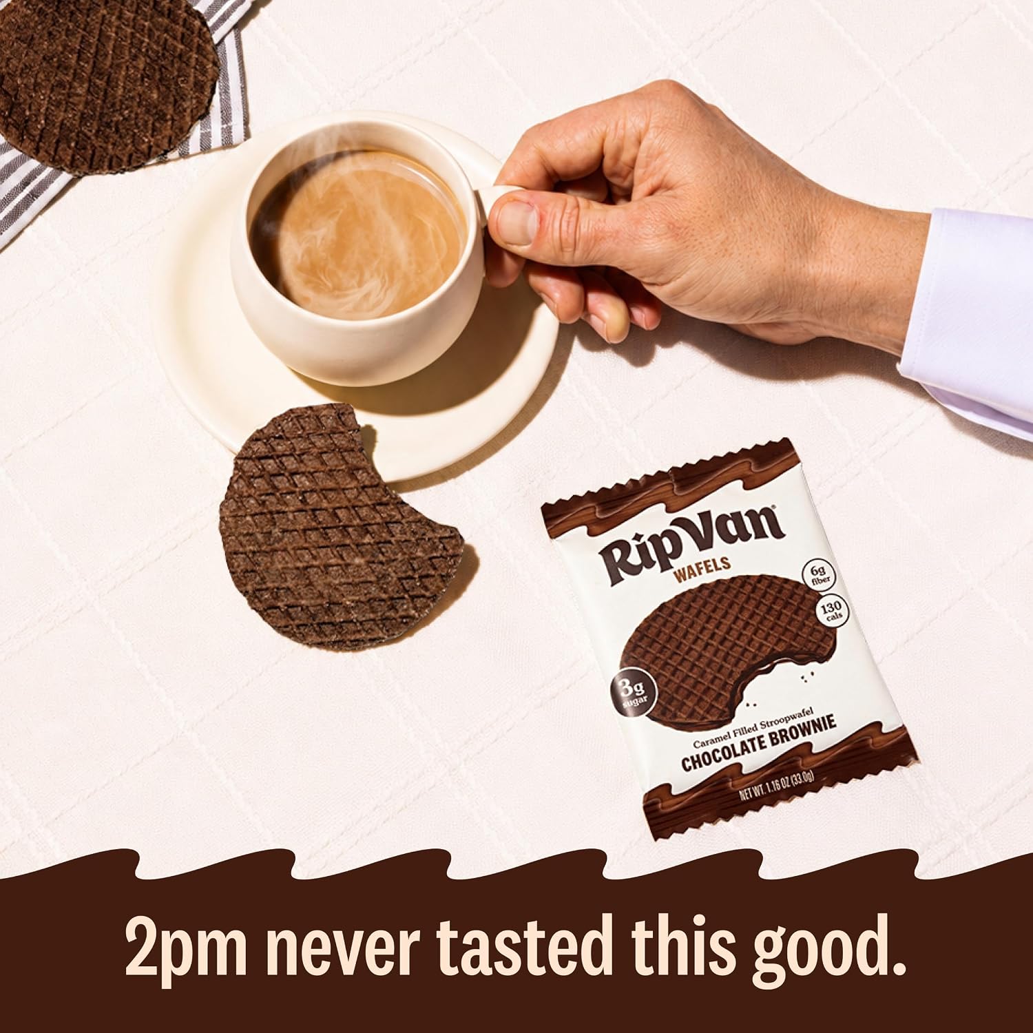A hand holds a steaming cup of coffee beside a chocolate brownie wafer, with a partially eaten wafer and a package of RipVan Wafels on a white tablecloth, promoting an afternoon treat.