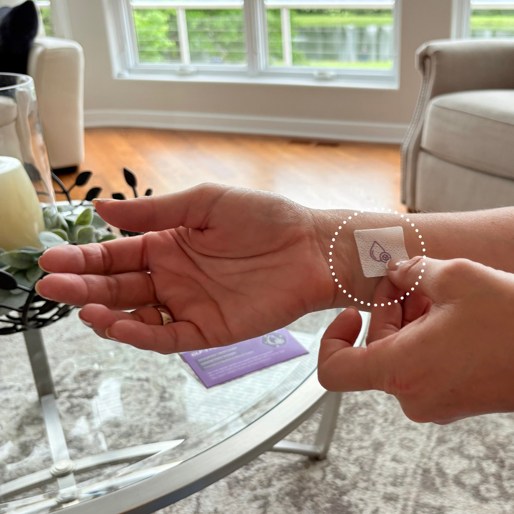 A person is applying a small, square patch to their wrist while seated in a cozy living room with a glass coffee table and natural light coming through the windows.
