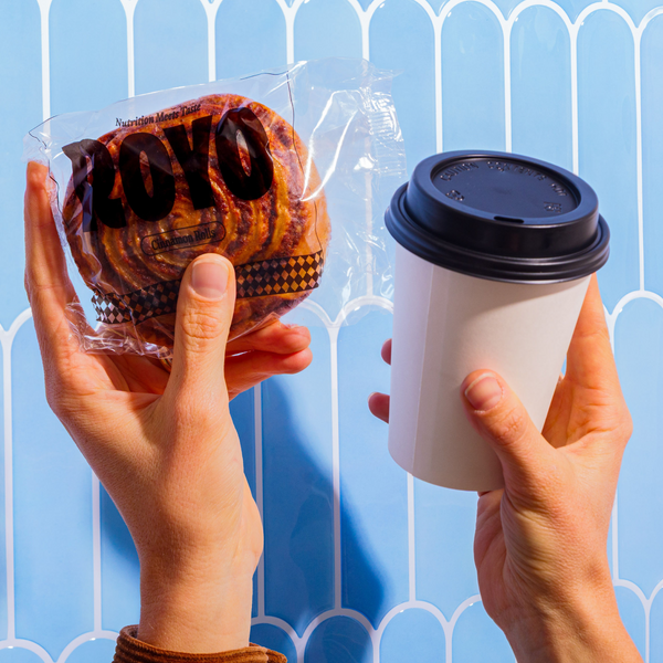 A person holds a packaged cinnamon roll in one hand and a white coffee cup in the other, set against a light blue background with a decorative pattern.