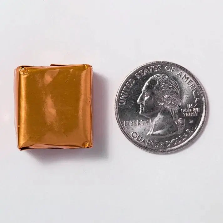 A shiny, gold-wrapped square candy is placed next to a U.S. quarter, highlighting its small size against the coin on a plain white background.