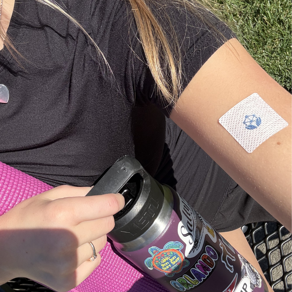 A person in a black shirt sits on a yoga mat, holding a water bottle with stickers, while a bandage with a blue symbol is visible on their arm.