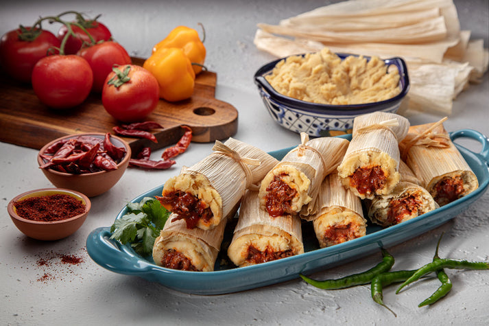 A blue serving dish holds several tamales filled with red sauce, garnished with cilantro, alongside fresh tomatoes, yellow peppers, dried chiles, and a bowl of hummus on a wooden cutting board.