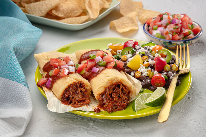 Two tamales filled with shredded meat, topped with diced tomatoes and onions, served on a green plate with a side of black bean salad, lime wedge, and tortilla chips in the background.