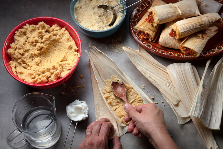 A person is preparing tamales using corn masa in a red bowl, with a spoon, while assembling them in corn husks on a gray surface, alongside a blue bowl of masa and measuring cups of water and lard.