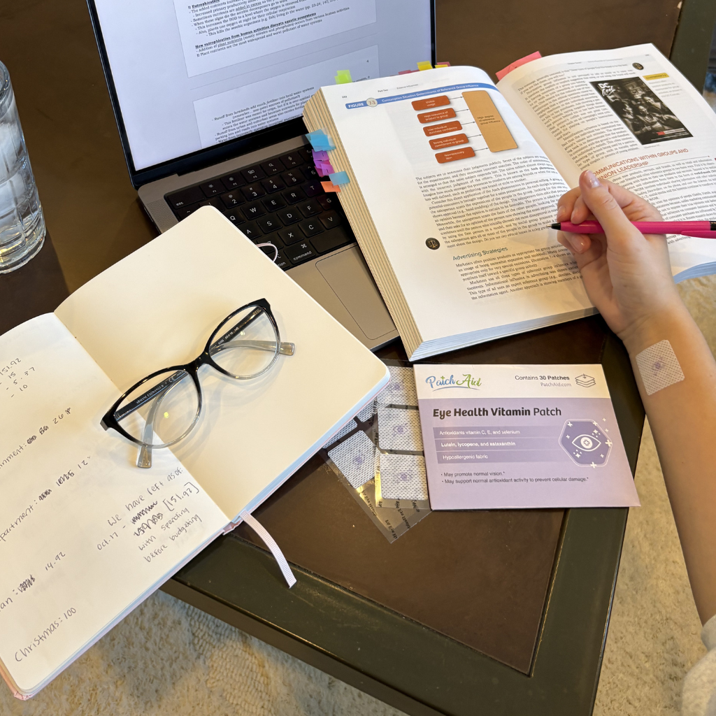 A hand holding a pink pen writes in an open notebook next to a laptop and an open book, while a glasses case and a packaging for Eye Health Vitamin Patches are also visible on a wooden table.