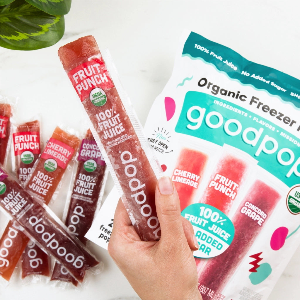 A hand holds a Fruit Punch organic freezer pop in front of a colorful package labeled "goodpop," with additional pops in the background showcasing flavors like Cherry Limeade and Concord Grape.