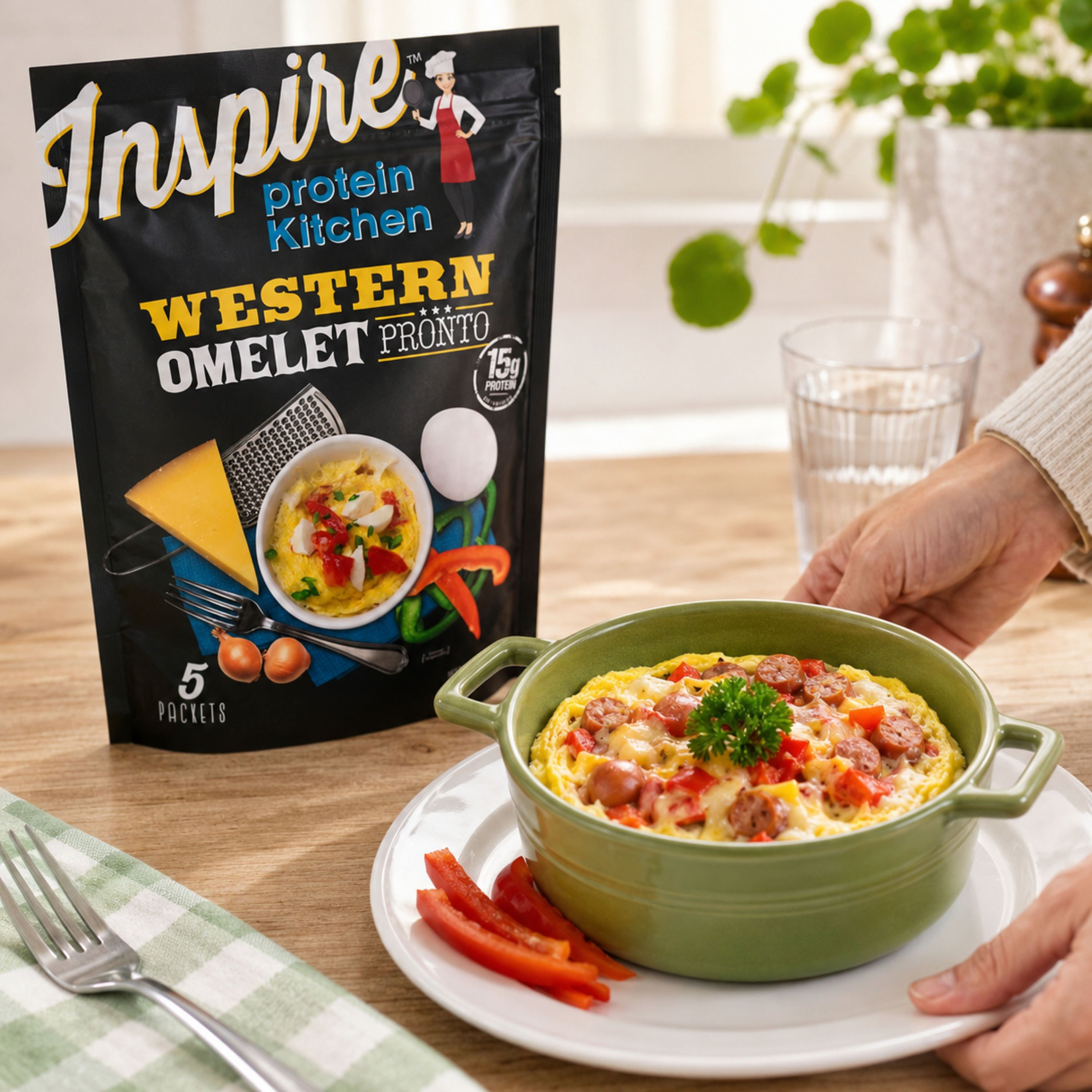 A person places a green dish of a colorful Western omelet, garnished with parsley, on a white plate beside sliced red peppers, with a packet of Inspire protein kitchen mix in the background on a wooden table.