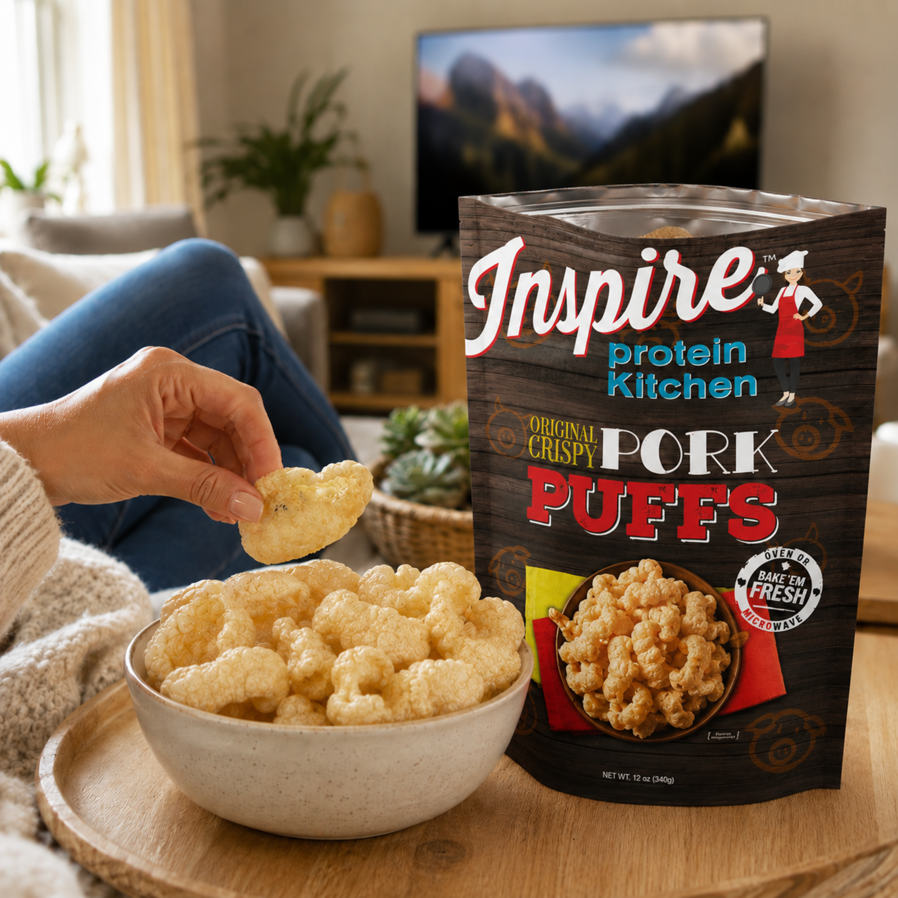 A person's hand holds a piece of crispy pork puffs above a bowl filled with the snack, next to a bag of Inspire Protein Pork Puffs.