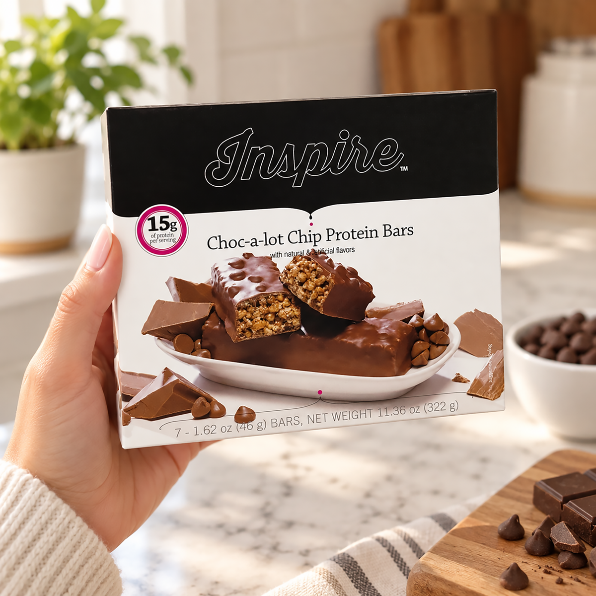 A hand holds a box of Inspire Choc-a-lot Chip Protein Bars, showcasing the product with chocolate-covered bars and chocolate chips on a kitchen counter with soft lighting.