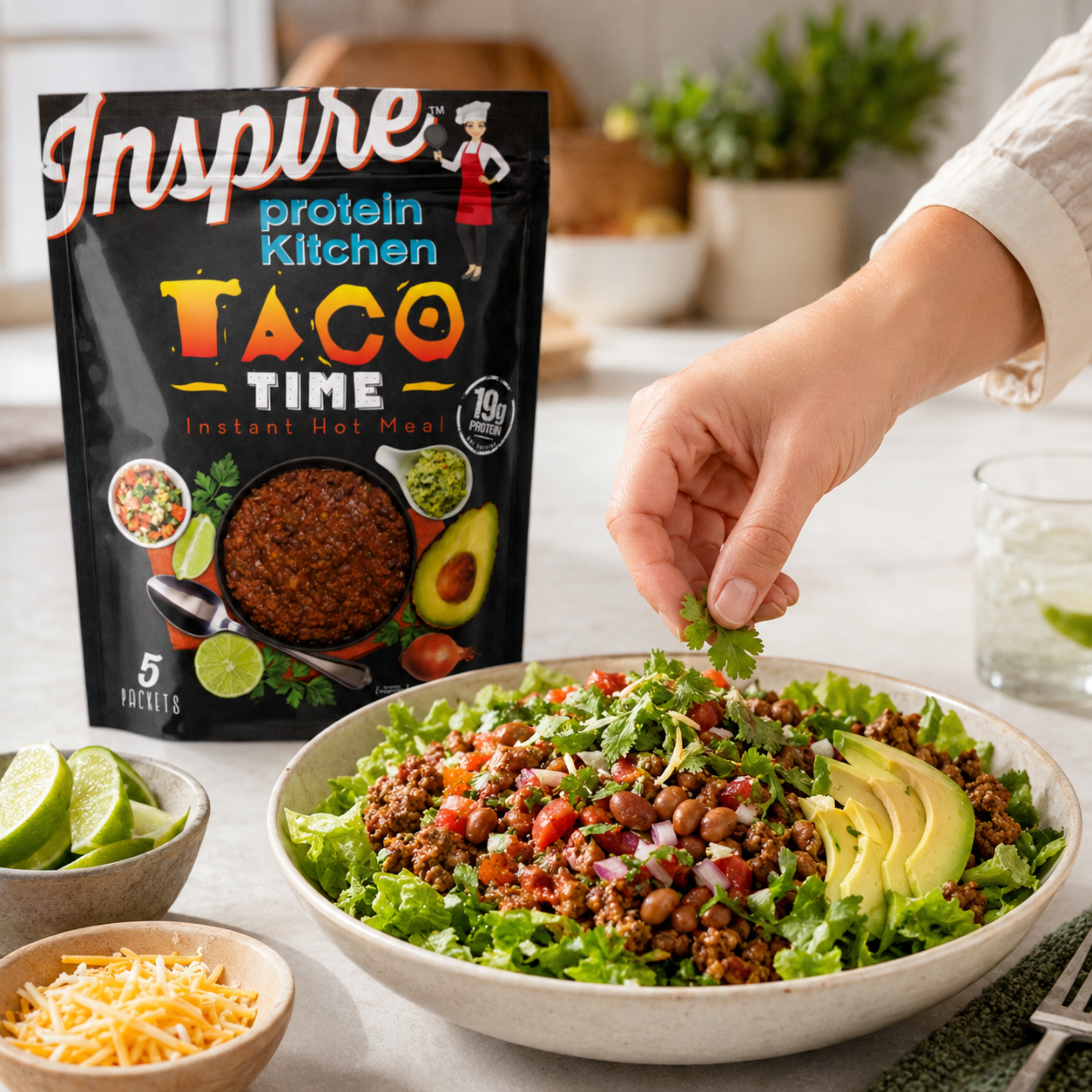 Hand adding cilantro to a taco salad with avocado, beans, tomatoes, and seasoned taco meat. Inspire Protein Kitchen Taco Time package is in the background.