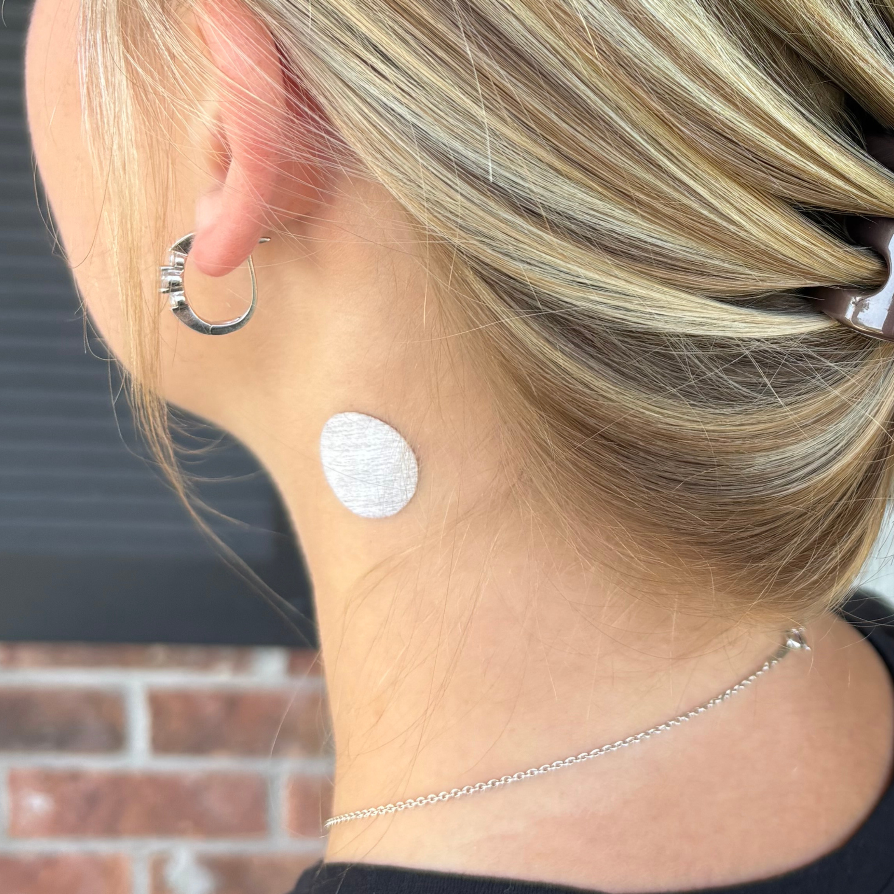 A person with long, blonde hair is shown from the side, revealing a round, white patch applied to the back of their neck, with a blurred brick wall in the background.