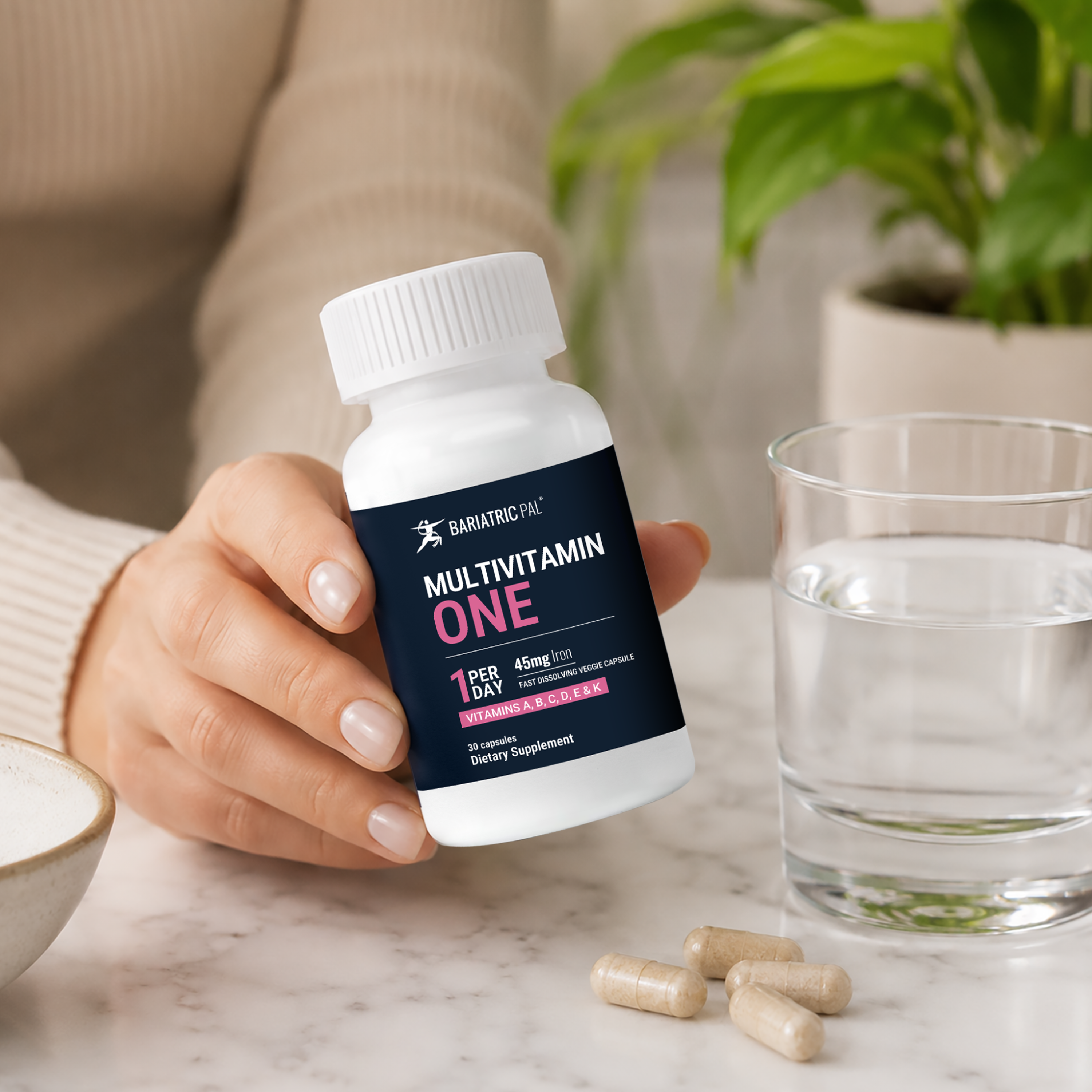 A hand holds a white bottle of BariatricPal Multivitamin One capsules, with four capsules resting on a marble surface next to a glass of water and a potted plant in the background.