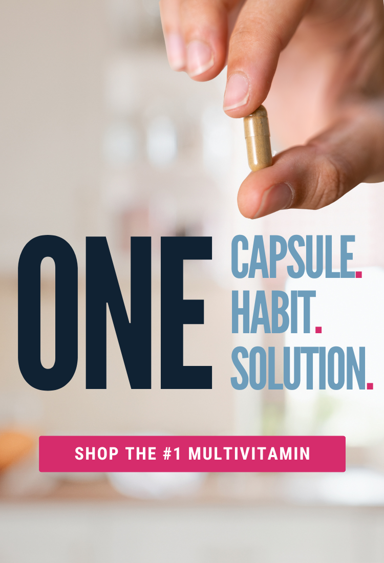 A hand holds a gold capsule against a blurred kitchen background, emphasizing the message "One Capsule. Habit. Solution." with a call to action to shop the number one multivitamin.