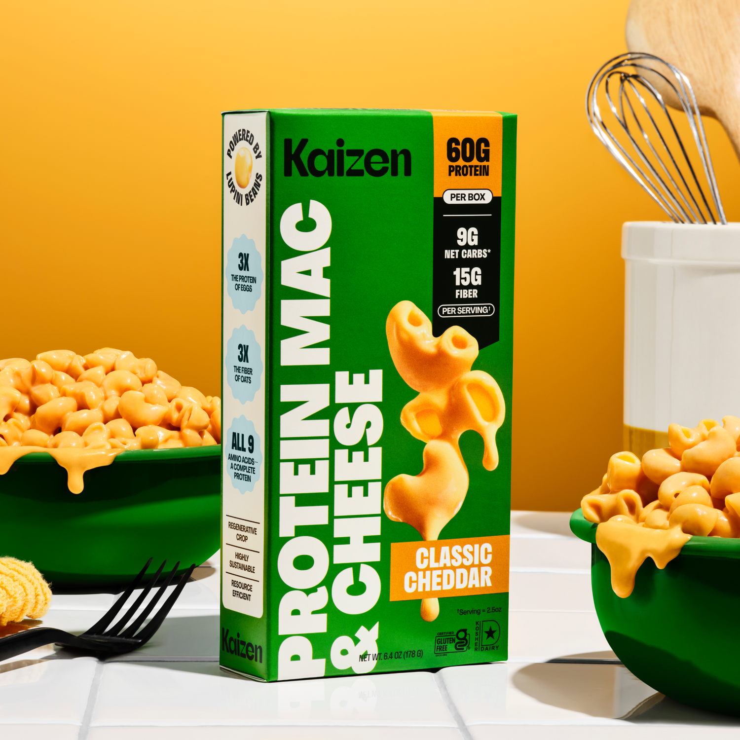 A bright yellow background features a box of Kaizen Protein Mac & Cheese with a green bowl of macaroni and cheese beside it, highlighting its nutritional benefits like high protein and fiber content.