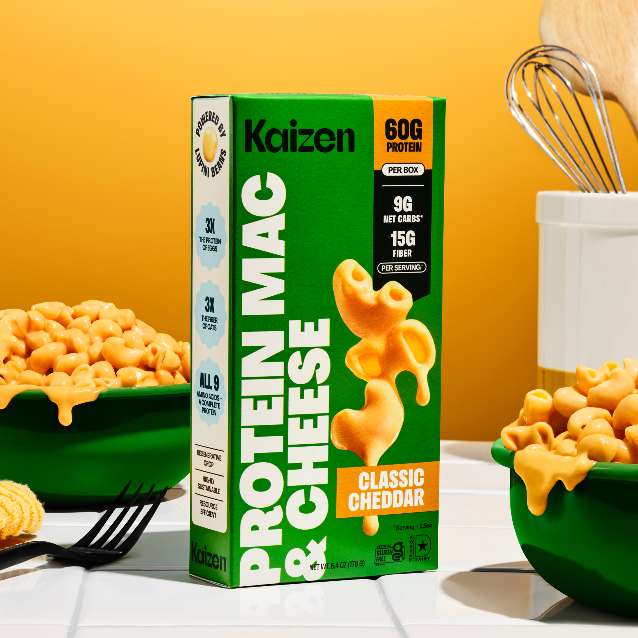 A green box of Kaizen Protein Mac & Cheese with classic cheddar flavor is displayed next to a bowl of creamy macaroni, set against a bright orange background with a whisk in the background.