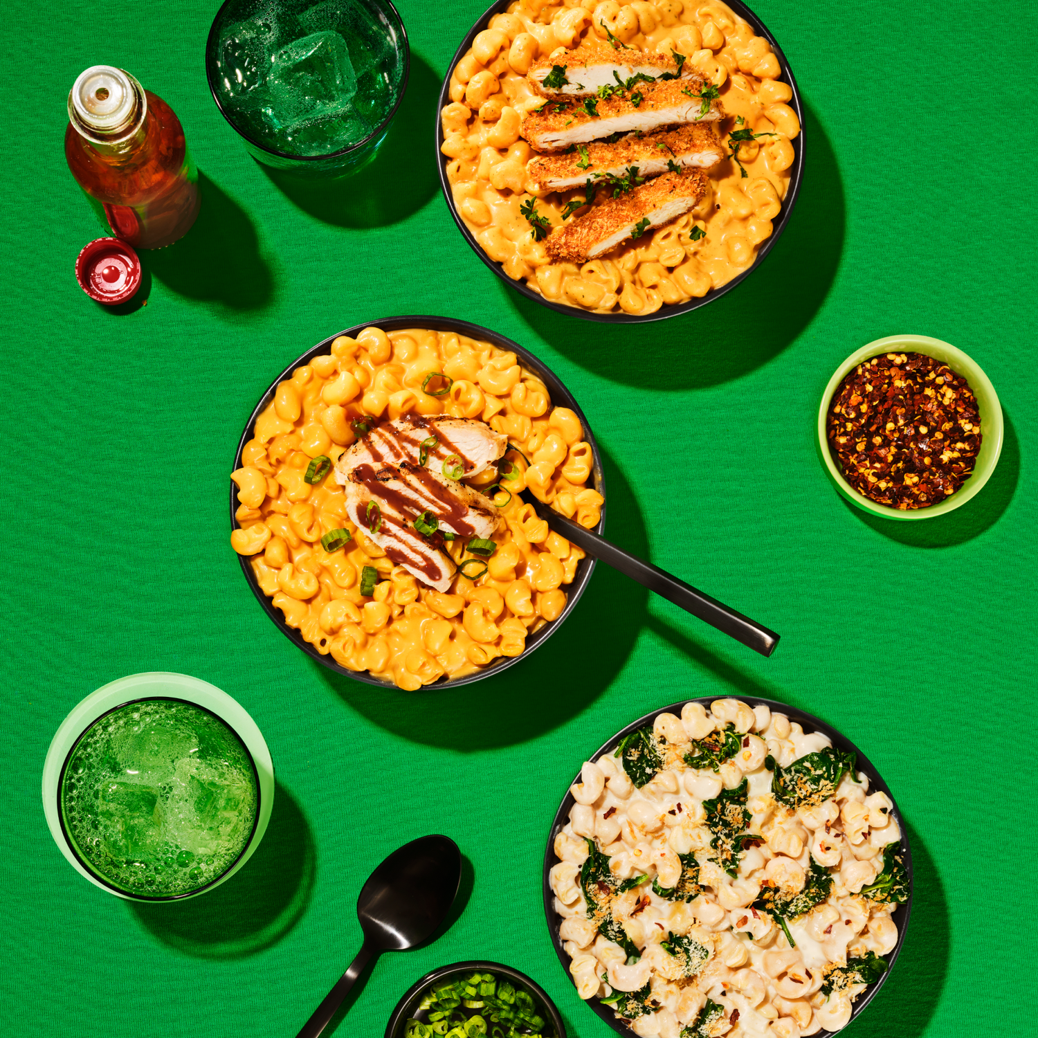 Three bowls of pasta with different sauces and toppings are arranged on a bright green surface, accompanied by a glass of green drink, a small bowl of chili flakes, and sliced green onions.