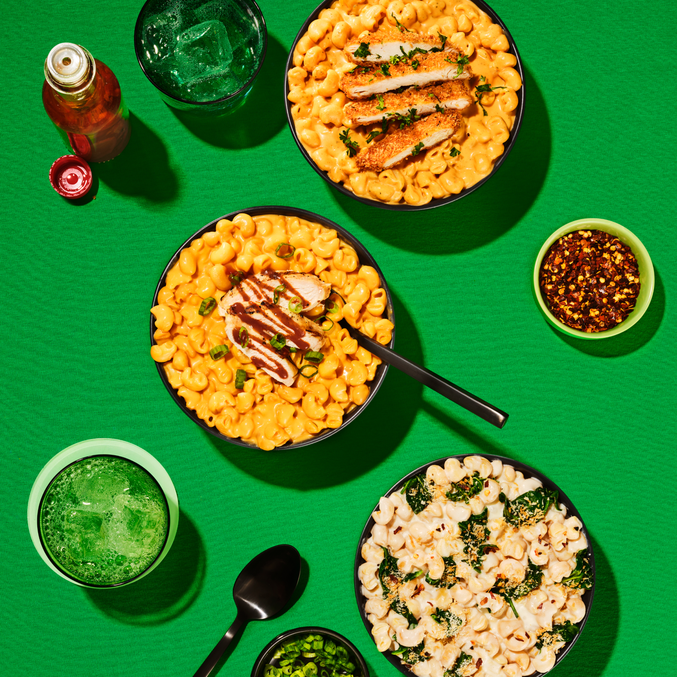 Three bowls of pasta dishes sit on a bright green table, featuring creamy mac and cheese with grilled chicken, a spinach and cheese mix, and a side of red pepper flakes, alongside drinks and utensils.