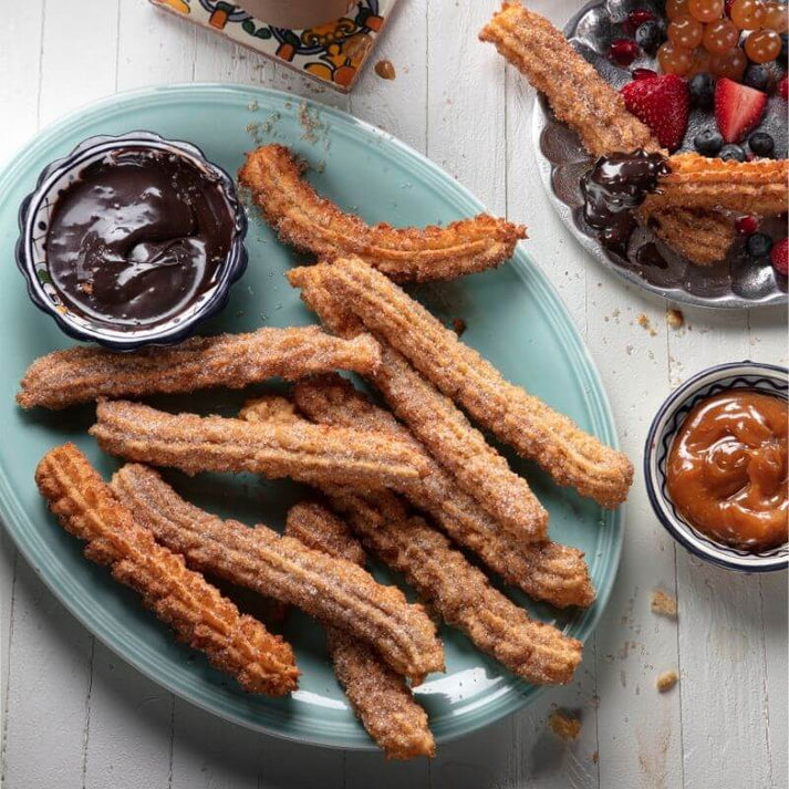 Crispy churros coated in cinnamon sugar on a blue platter, served with chocolate and caramel dipping sauces, alongside a bowl of mixed berries.