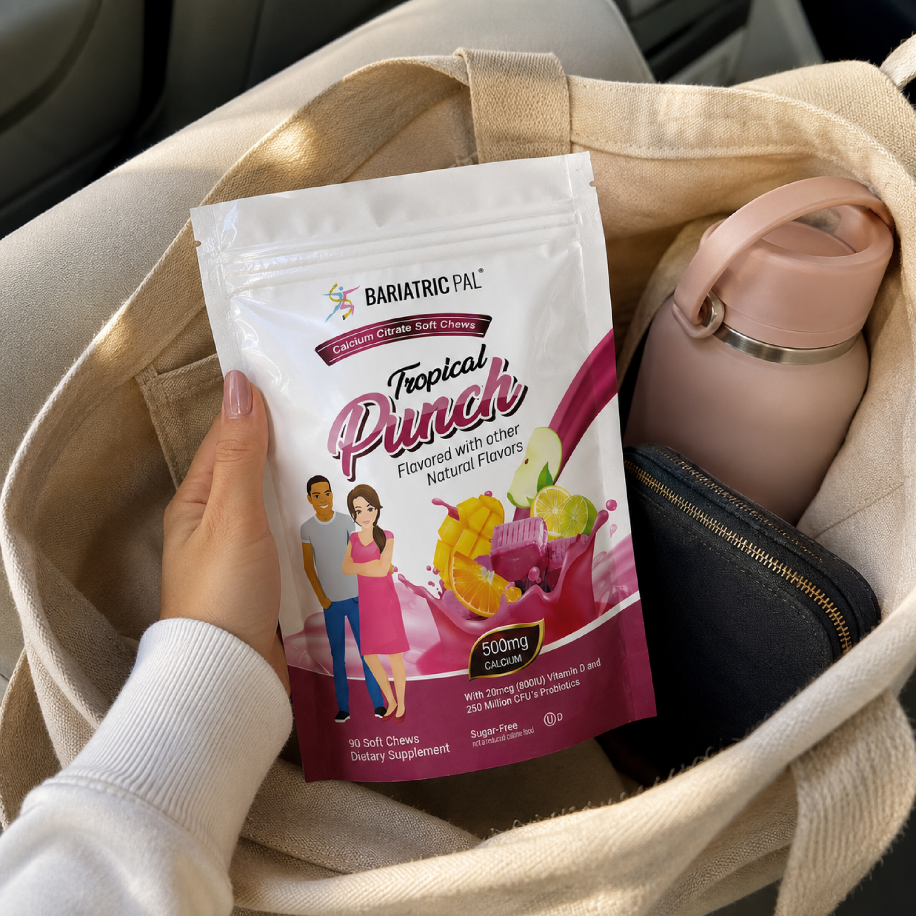 A hand holds a stand-up pouch of BariatricPal Tropical Punch dietary supplement inside a beige tote bag, next to a pink water bottle and a black wallet.