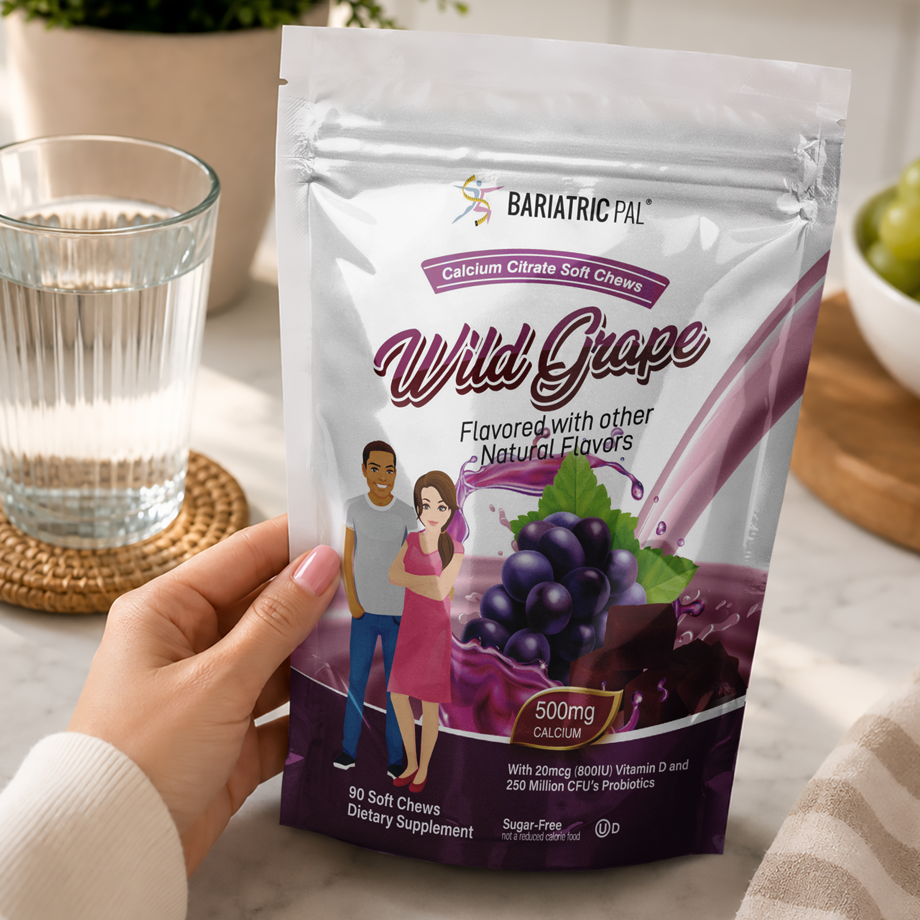 A hand holds a stand-up pouch of BariatricPal Wild Grape Calcium Citrate Soft Chews. The packaging is silver and purple with illustrations of grapes and a couple, set against a blurred kitchen background with a glass of water.