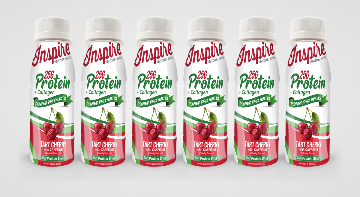 Six bottles of Inspire Protein Shots are lined up, each labeled with "25G Protein + Collagen" and "Tart Cherry with Caffeine," featuring a red and green design with cherries on the front.