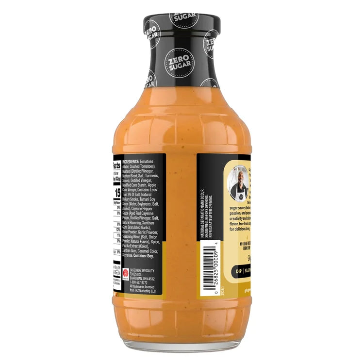 A clear glass bottle filled with a creamy orange sauce features a black cap, a label listing ingredients, and a photo of a chef, indicating it is a zero-sugar product suitable for dipping or dressing.
