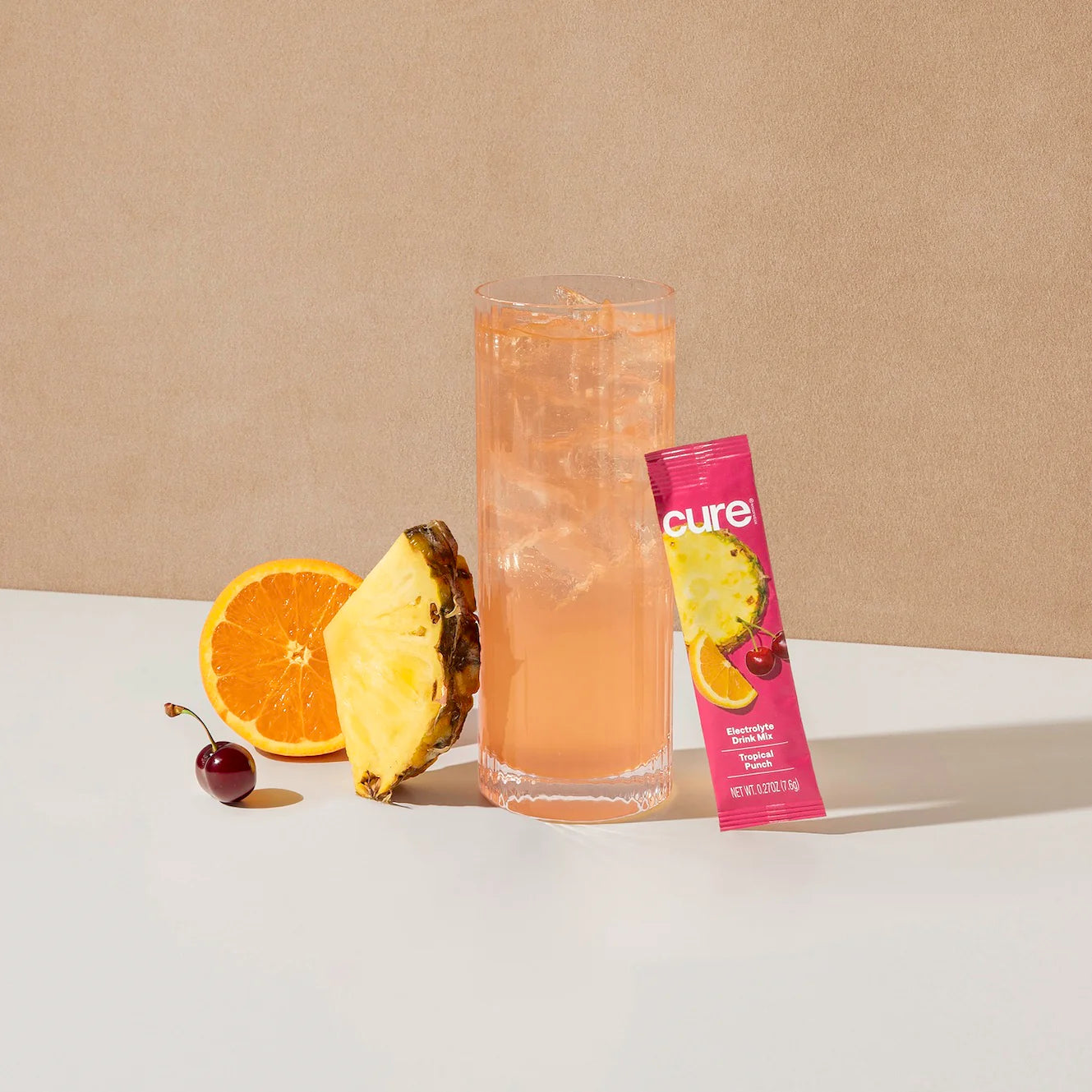 A tall glass of pink beverage with ice is surrounded by a slice of orange, a piece of pineapple, a cherry, and a packet of Cure electrolyte drink mix on a light surface against a beige background.
