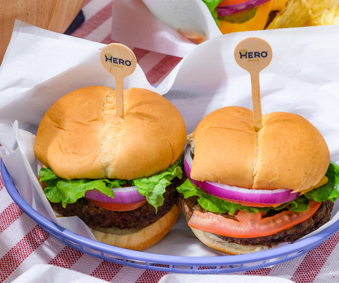 Two freshly made burgers with lettuce, tomato, and red onion are served in a blue basket, each topped with a wooden pick labeled "HERO," on a red and white checkered tablecloth.