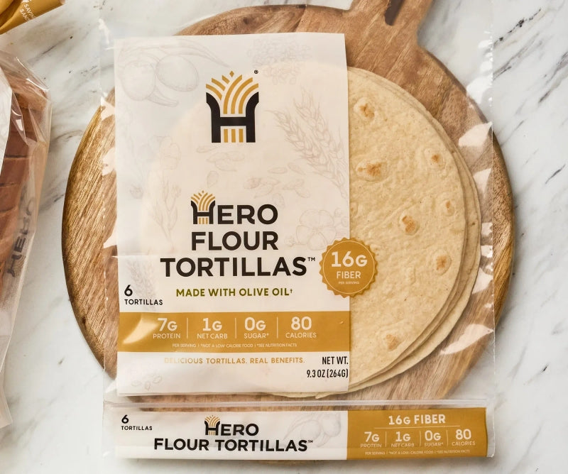 A package of Hero Flour Tortillas, featuring six tortillas made with olive oil, is displayed on a wooden cutting board, highlighting nutritional information and benefits on the packaging.