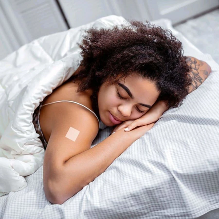 A young woman with curly hair peacefully sleeps on a bed, covered by a white blanket, with a small bandage on her arm, suggesting recent medical care or a vaccination.